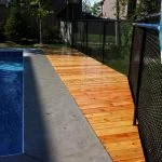 a wooden pool deck crafted by stittsville deck builder extends along the side of a blue in-ground swimming pool, bordered by a black mesh fence, with houses and trees visible in the background on a