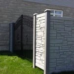 a black wrought iron gate is set within a gray stone wall, adjacent to a white simtek fence installation, under a clear sky on a sunny day.