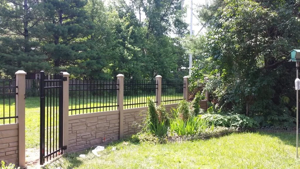 a simtek fence with stone pillars borders a lush green yard with trees and a variety of plants on a sunny day, farm fence