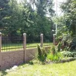 a simtek fence with stone pillars borders a lush green yard with trees and a variety of plants on a sunny day.