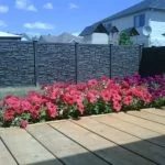 a wooden deck overlooks a vibrant flowerbed against a dark simtek fence under a clear sky, offering the benefits of durability and privacy.