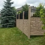 a stone and iron fence, showcasing the benefits of using a simtek fence, borders a neatly trimmed lawn with trees and a residential building in the background on a cloudy day. a discarded water bottle lies