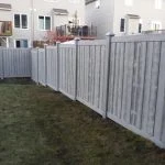 a simtek fence encloses a backyard with a grassy lawn and the rear facade of a residential building, featuring a deck and stairs, is visible in the background.