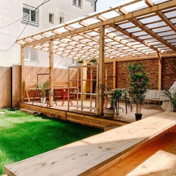 a well-lit wooden patio area, building a pergola, furnished with chairs and a table, surrounded by green plants and enclosed by a brick wall and wooden fence.