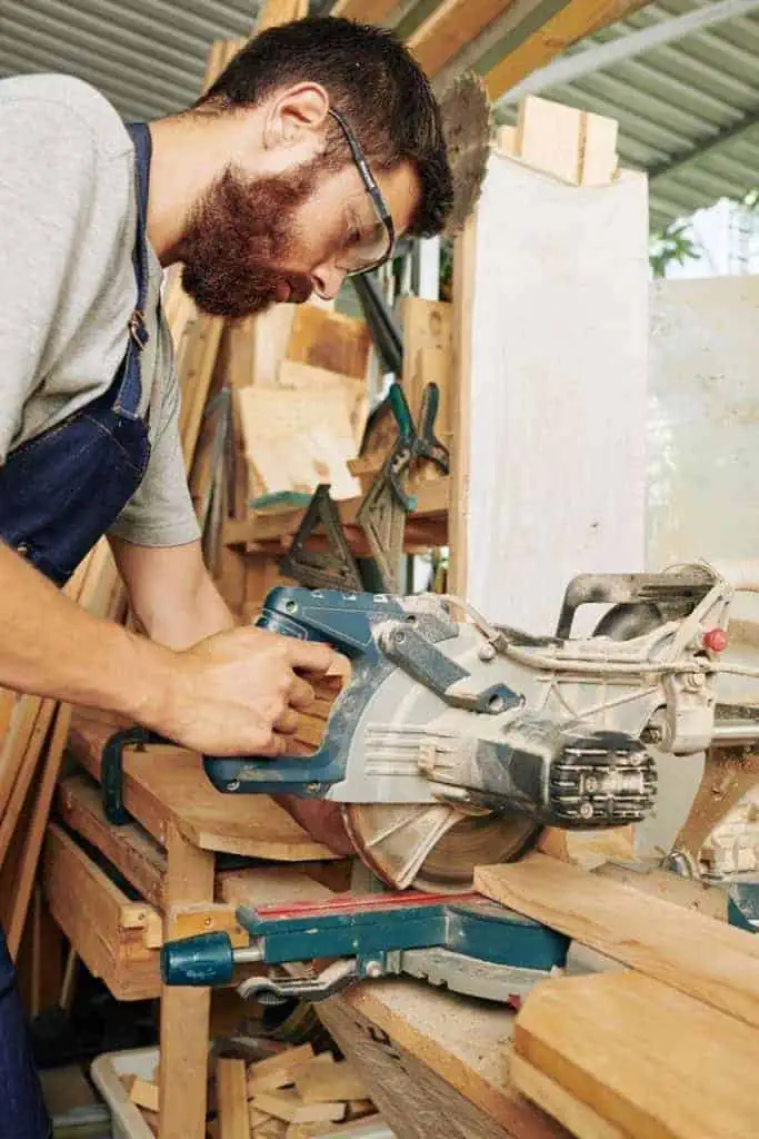 a bearded man in overalls using an electric saw to cut a piece of wood in a woodworking shop.