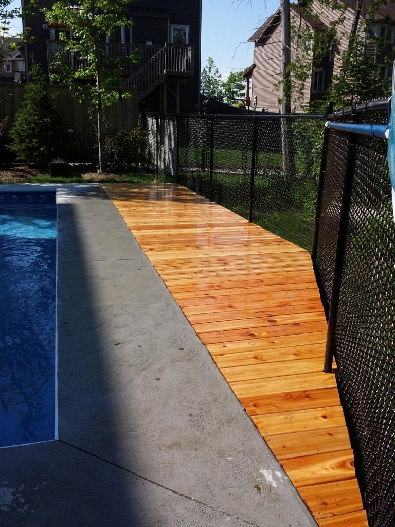 A sunlit wooden walkway beside a pool, accented by sleek chain-link fence installation in Ottawa, with trees and houses forming a serene backdrop.