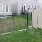 a residential backyard benefitting from a chain-link fence adjoining a beige vinyl privacy fence, with houses featuring decks in the background.