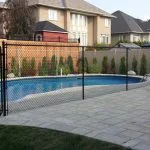 a person is seen from behind looking out over a residential backyard featuring a patio, a swimming pool with a chain-link fence around it, highlighting the benefits of such fencing, and the back exteriors of