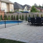 A residential backyard featuring a swimming pool with a chain-link safety fence around it and a dining area with a table and chairs on a stone patio.