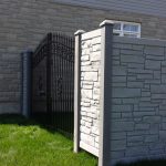 A black wrought iron gate is set within a gray stone wall, adjacent to a white SimTek fence installation, under a clear sky on a sunny day.