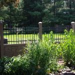 A garden with tall green plants near a SimTek fence and brick columns, with trees and a lawn in the background on a sunny day.