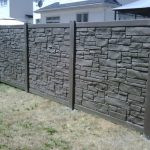 A section of a stone-textured SimTek fence stands on grass, with part of a residential structure visible in the background.