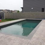 A rectangular inground swimming pool enclosed by a stone wall and a simtek fence, with a trampoline in the corner of a residential backyard.