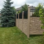 A stone and iron fence, showcasing the benefits of using a SimTek fence, borders a neatly trimmed lawn with trees and a residential building in the background on a cloudy day. A discarded water bottle lies