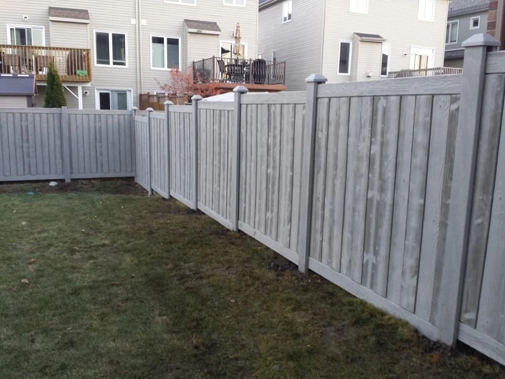 a simtek fence encloses a backyard with a grassy lawn and the rear facade of a residential building, featuring a deck and stairs, is visible in the background.