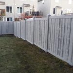 A Simtek fence encloses a backyard with a grassy lawn and the rear facade of a residential building, featuring a deck and stairs, is visible in the background.