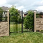 A black metal gate stands closed, set between two decorative stone pillars that highlight the benefits of using a SimTek fence, with a well-maintained lawn and residential structures in the background.