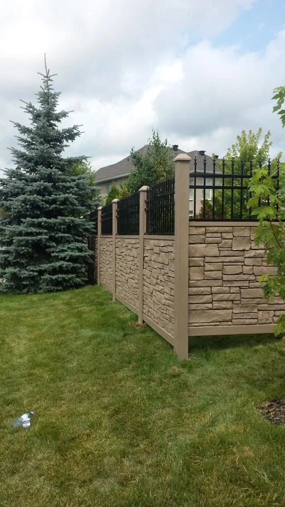 a stone and metal fence, reminiscent of the quality found in simtek fence installations in ottawa, runs alongside a green lawn with a large evergreen tree and a house in the background under a cloudy sky.