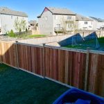 A cedar privacy fence separates two suburban backyards with grass lawns; one yard features a soccer net. Row houses and a clear blue sky are visible in the background.