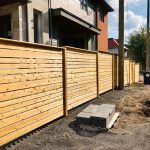 A new cedar wooden fence alongside a residential building on a sunny day, with construction materials and debris visible on the ground.