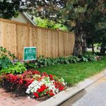A cedar fence lines a curving sidewalk with a small garden bed hosting a variety of plants and a sign indicating the entrance to a residential area.