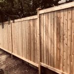 A cedar wooden fence with vertical planks follows a dirt path adjacent to a planted area with trees and shrubs.