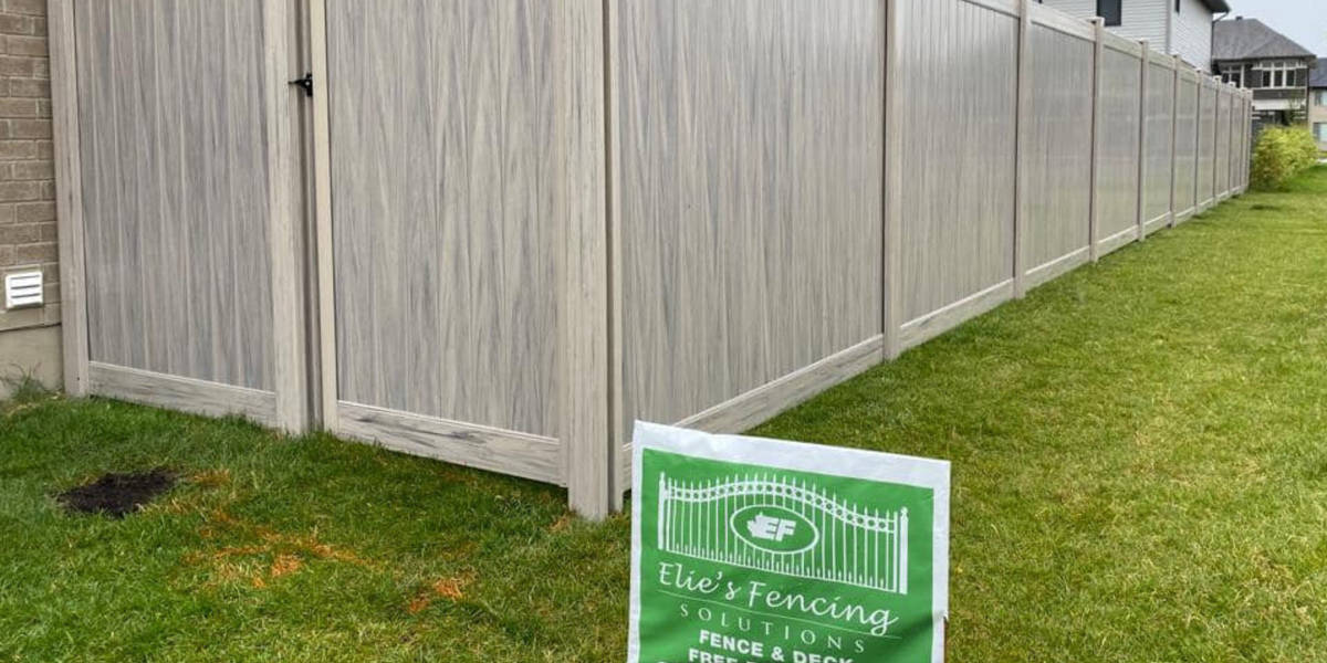 A light-colored privacy fence offers sturdy fence protection, separating a grassy yard from neighboring houses. A sign in the foreground advertises Elie's Fencing Solutions.