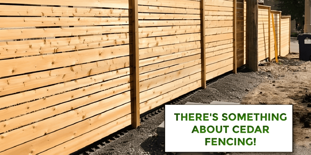 A long cedar privacy fence with vertical posts stands on a gravel surface. A sign in the foreground reads, "There's something about cedar fencing!.