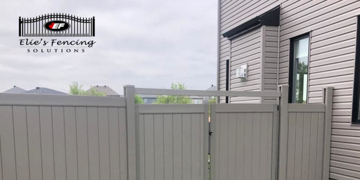 A tall, light gray privacy fence stands beside a modern house with siding, showcasing expert fence painting Ottawa homeowners trust. The Elie's Fencing Solutions logo appears in the top left corner.