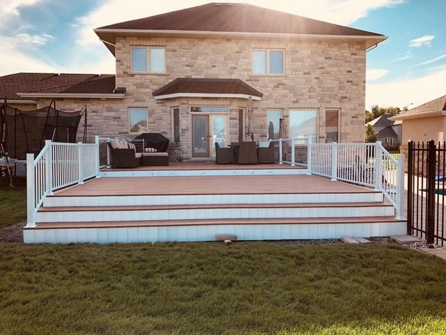 a residential backyard featuring a large deck with a white railing, patio furniture, and a two-story home in the background during daylight.