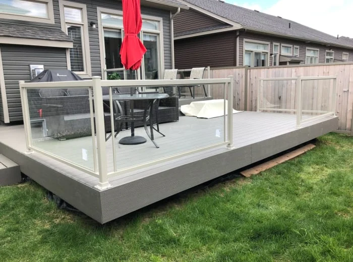 a residential backyard featuring a newly built, spacious deck with a glass railing, furnished with an outdoor dining table set and a grill under a closed red umbrella.