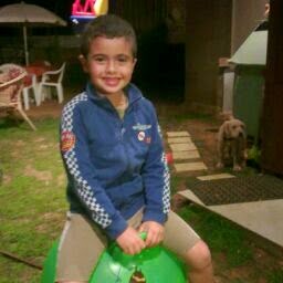 A young boy smiling for the camera while sitting on a green plastic toy in a backyard at dusk, with a dog visible in the background.