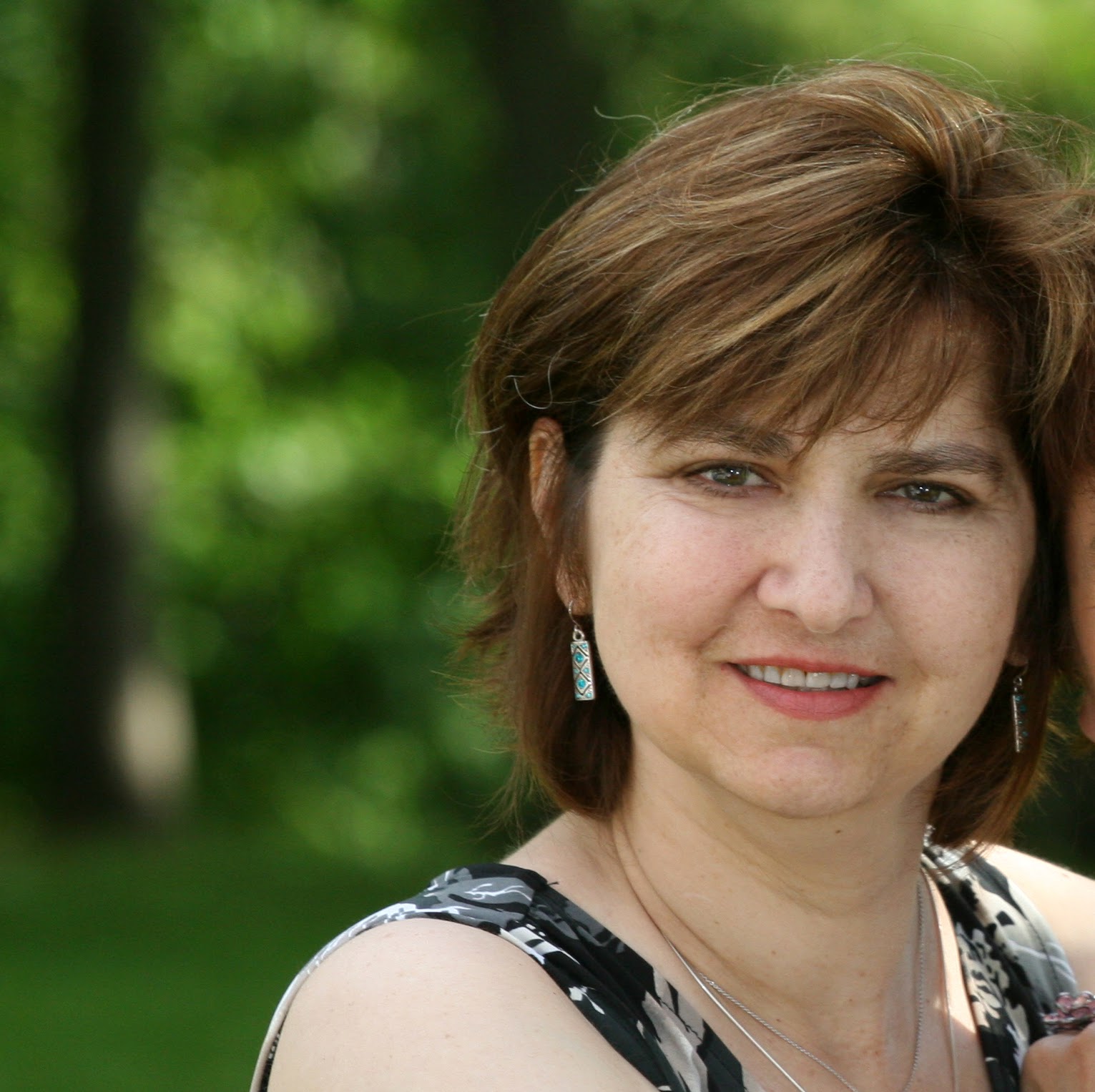 a middle-aged woman with shoulder-length brown hair and earrings, smiling slightly at the camera with a backdrop of green foliage.