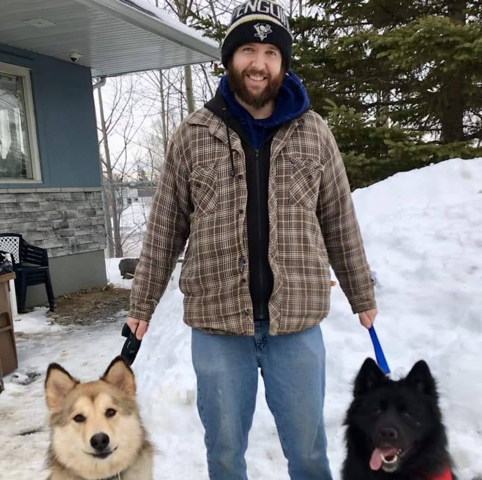 a man in winter clothing stands outdoors holding leashes of two dogs, with one dog having light fur and the other with dark fur, against a backdrop of a house and snow.