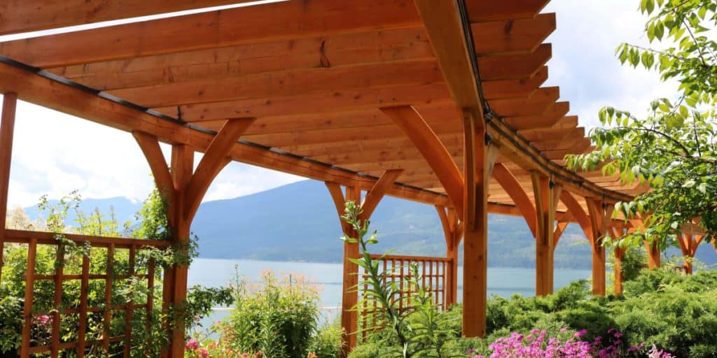 Expertly crafted by Pergola Builders Ottawa, this wooden pergola overlooks a serene lake with majestic mountains in the background, surrounded by lush greenery and vibrant flowering plants.