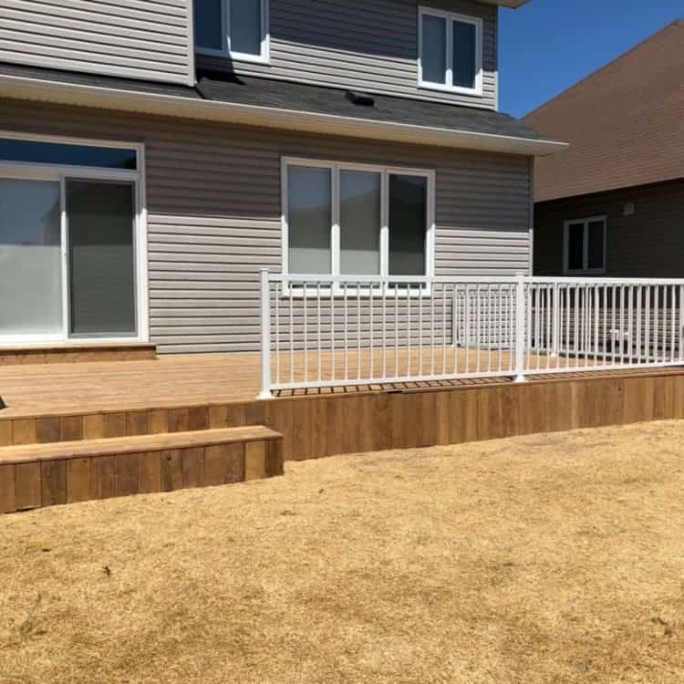 A newly built wooden deck by Ottawa deck builders with white railing in the backyard of a two-story house, featuring a few steps leading down to a lawn area covered with straw mulch, deck builders ottawa