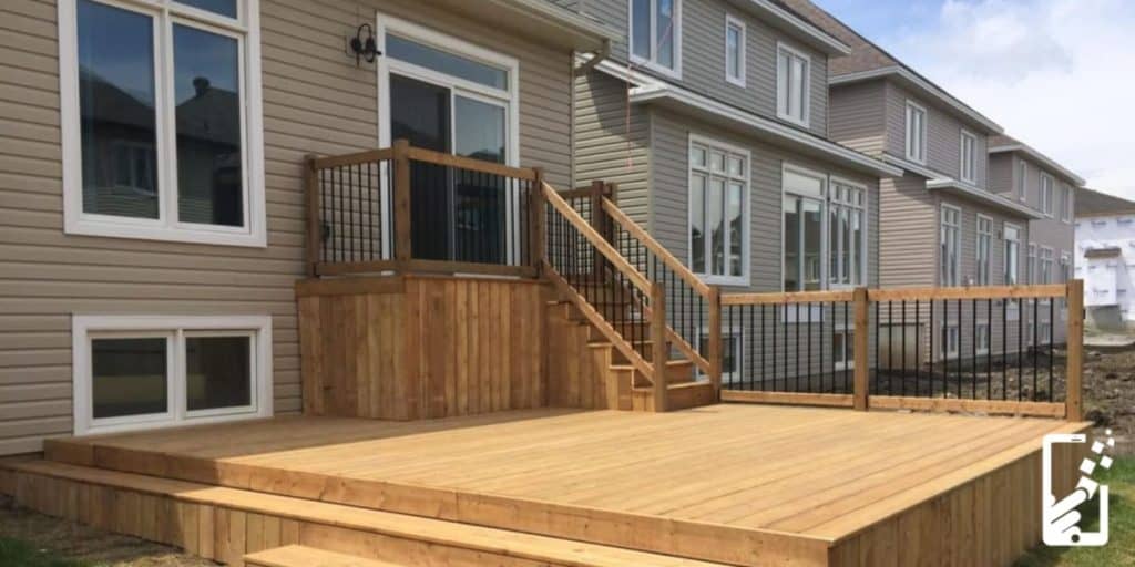 A wooden deck with stairs and railing, crafted by an Ottawa Deck Builder, is attached to the back of a beige house. Other similar homes are visible in the background.