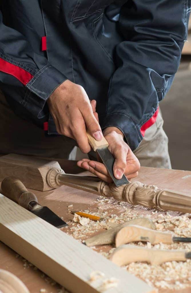 A craftsman is carefully using a chisel to shape a wooden object on a workbench, surrounded by wood shavings and a few other woodworking tools.