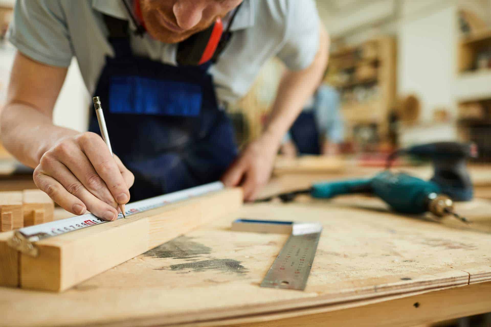 A person in a workshop measuring and marking wood with a ruler and pencil, with tools scattered around on the workbench, Dog fence installation, Fence Builder