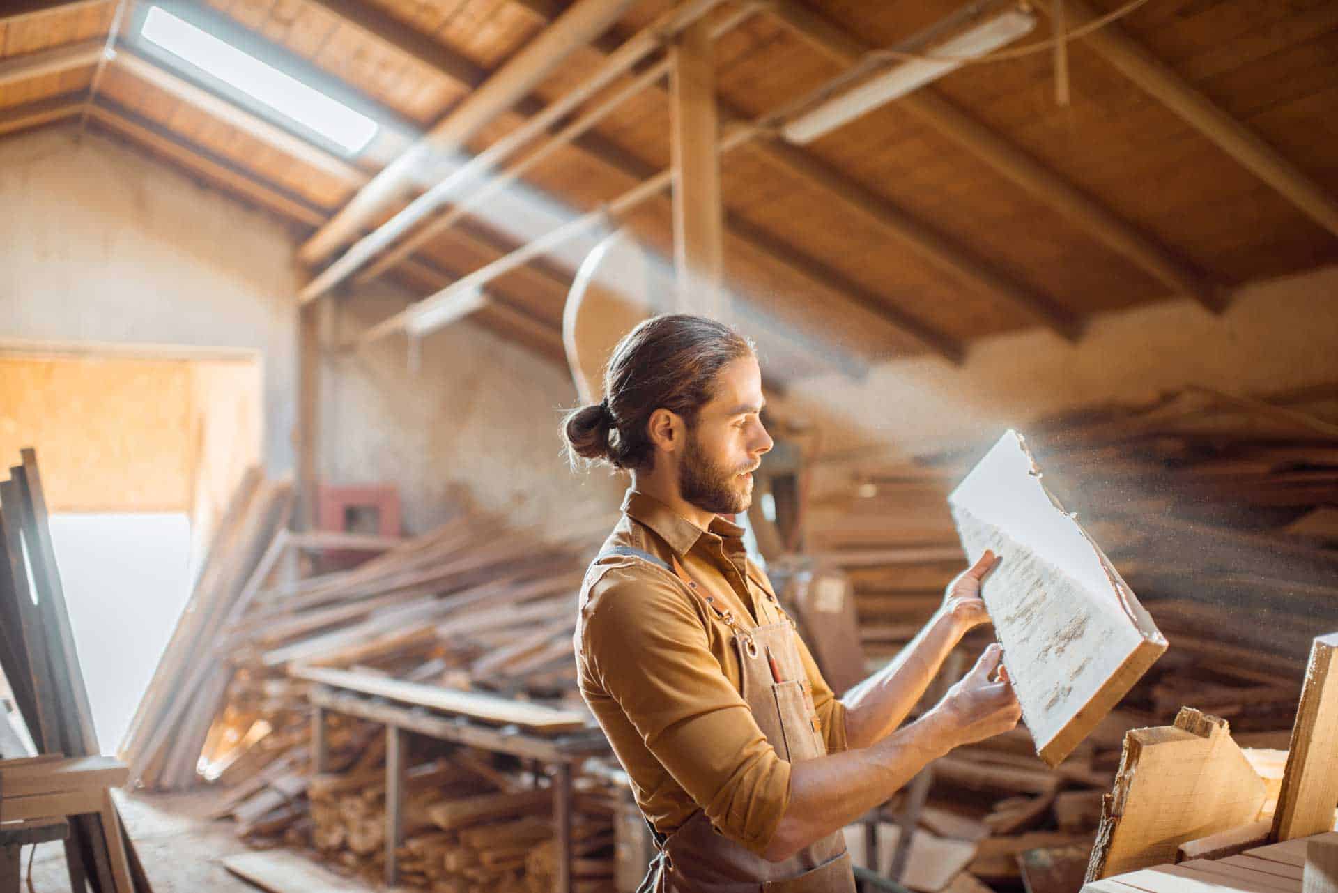 A man with a beard is examining a piece of wood in a sunlit woodworking shop.