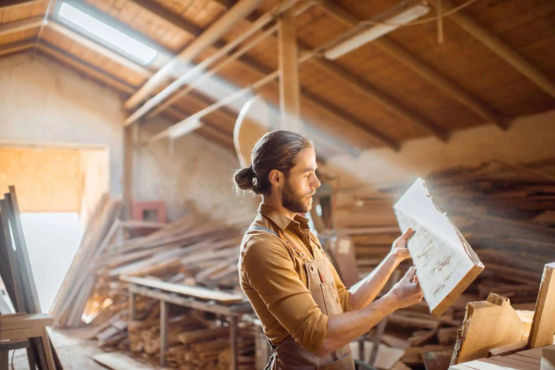 a man with a beard is examining a piece of wood in a sunlit woodworking shop.