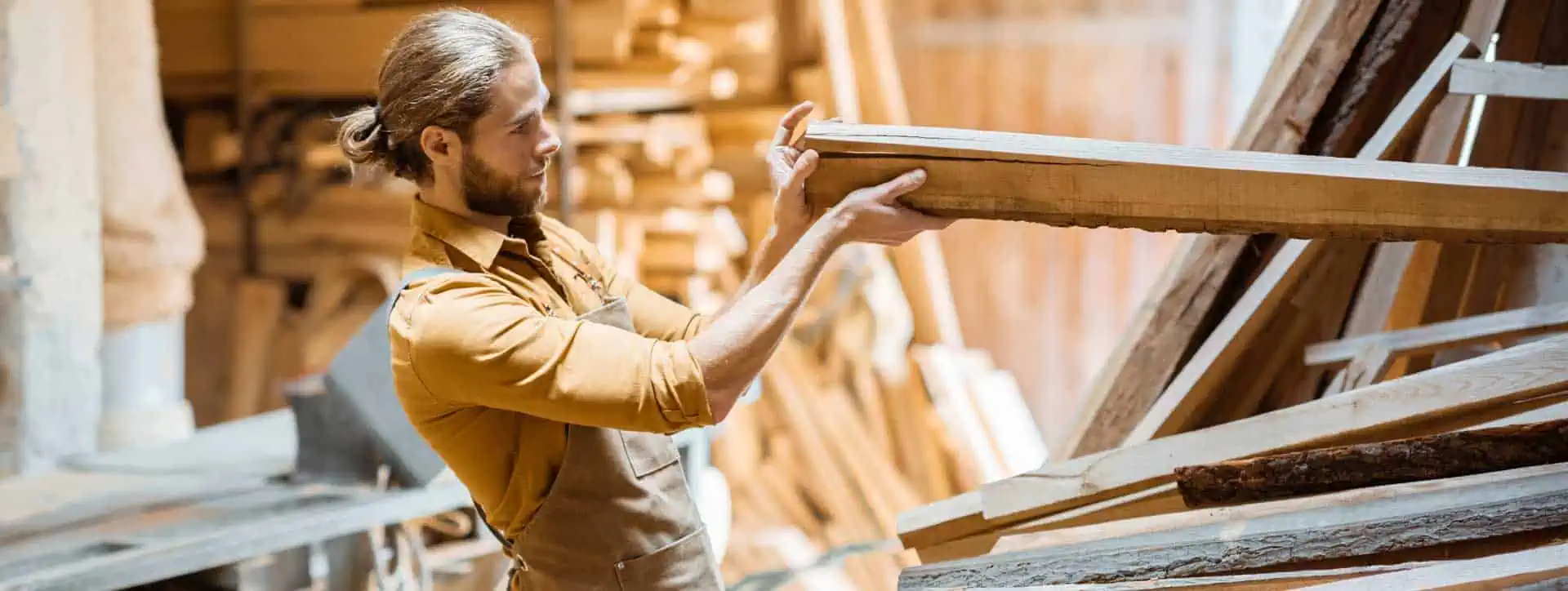 a man with a beard wearing an apron is examining a piece of lumber in a woodworking shop,dog fence installation, fence builder