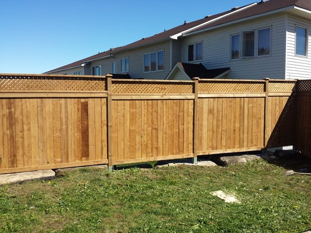 A newly constructed wooden fence stands in a backyard on a sunny day, with residential houses visible in the background.