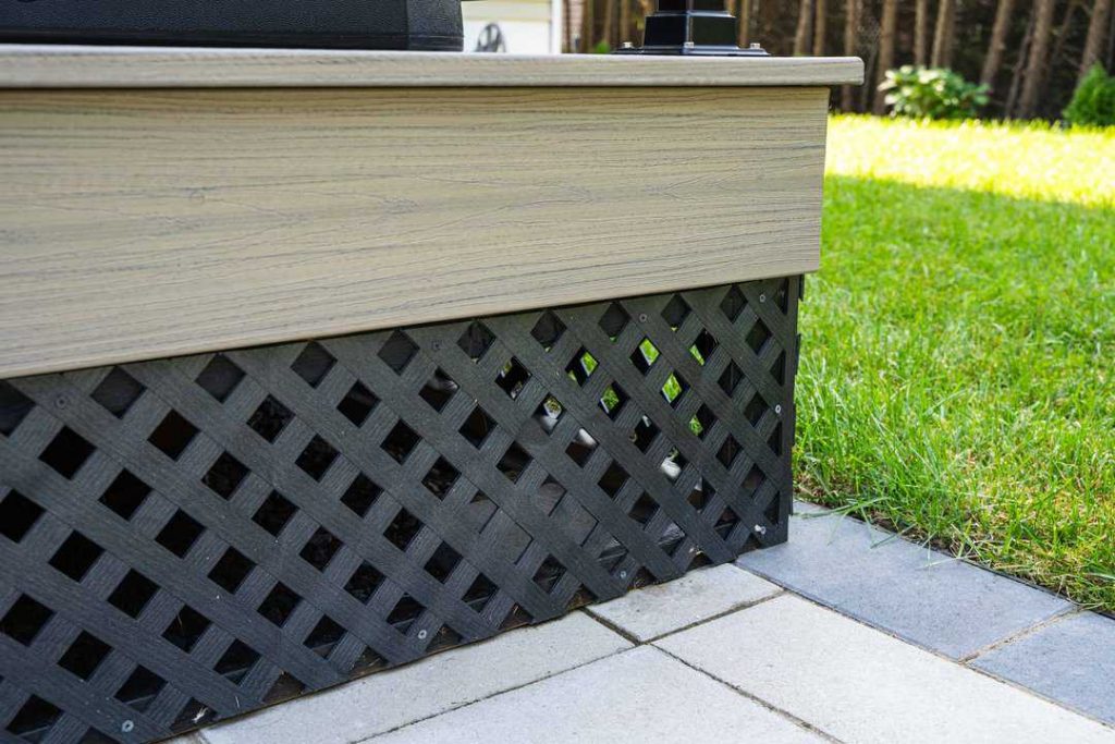The wooden deck edge features black lattice skirting, beautifully complementing the interlock paving of the Orleans-style tiled patio, all adjacent to a lush green lawn, Top-rated interlock paving Ottawa