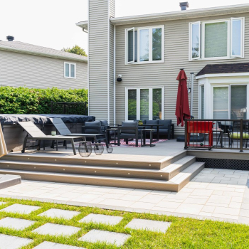A well-maintained backyard patio with outdoor furniture, featuring steps with built-in lighting leading up to a deck area adjacent to a two-story house.