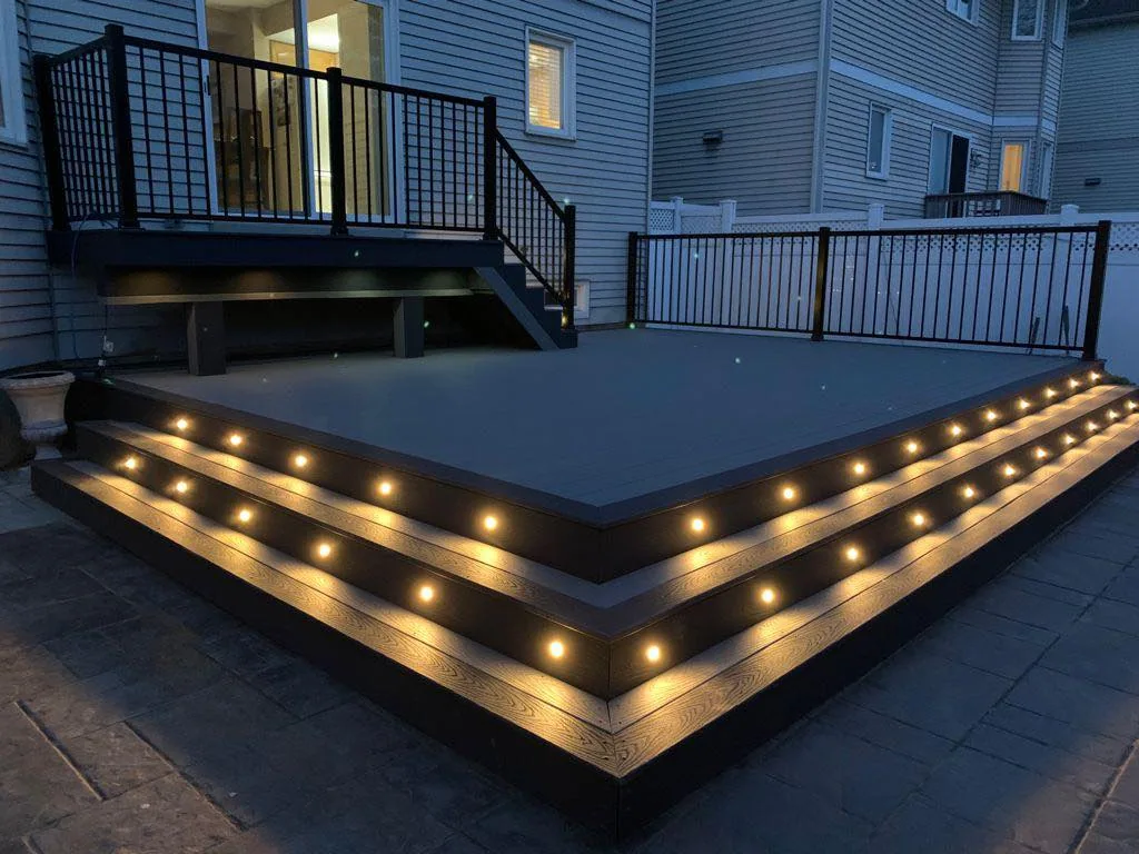 a residential backyard at dusk featuring a large composite deck with integrated lighting along the steps and railings, and an outdoor kitchen.