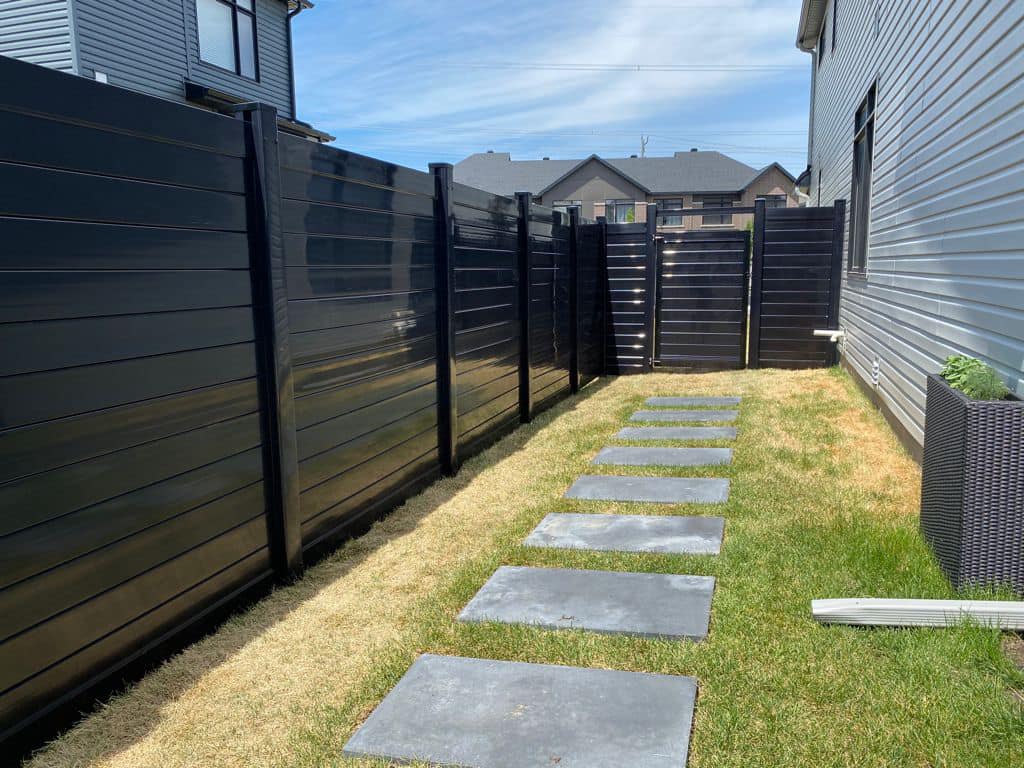 A narrow backyard pathway with rectangular stone slabs spaced evenly on grass, flanked by high black home fences and modern houses.