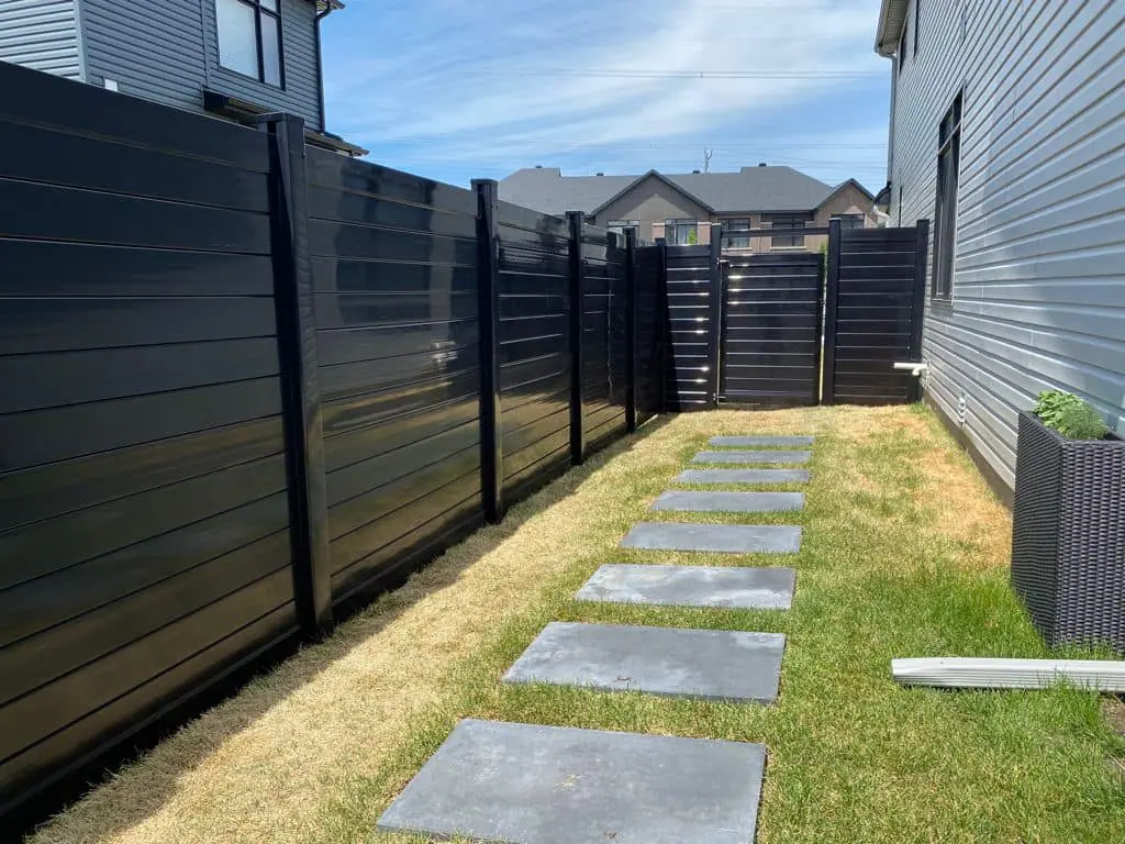 a narrow backyard pathway with rectangular stone slabs spaced evenly on grass, flanked by high black home fences and modern houses.