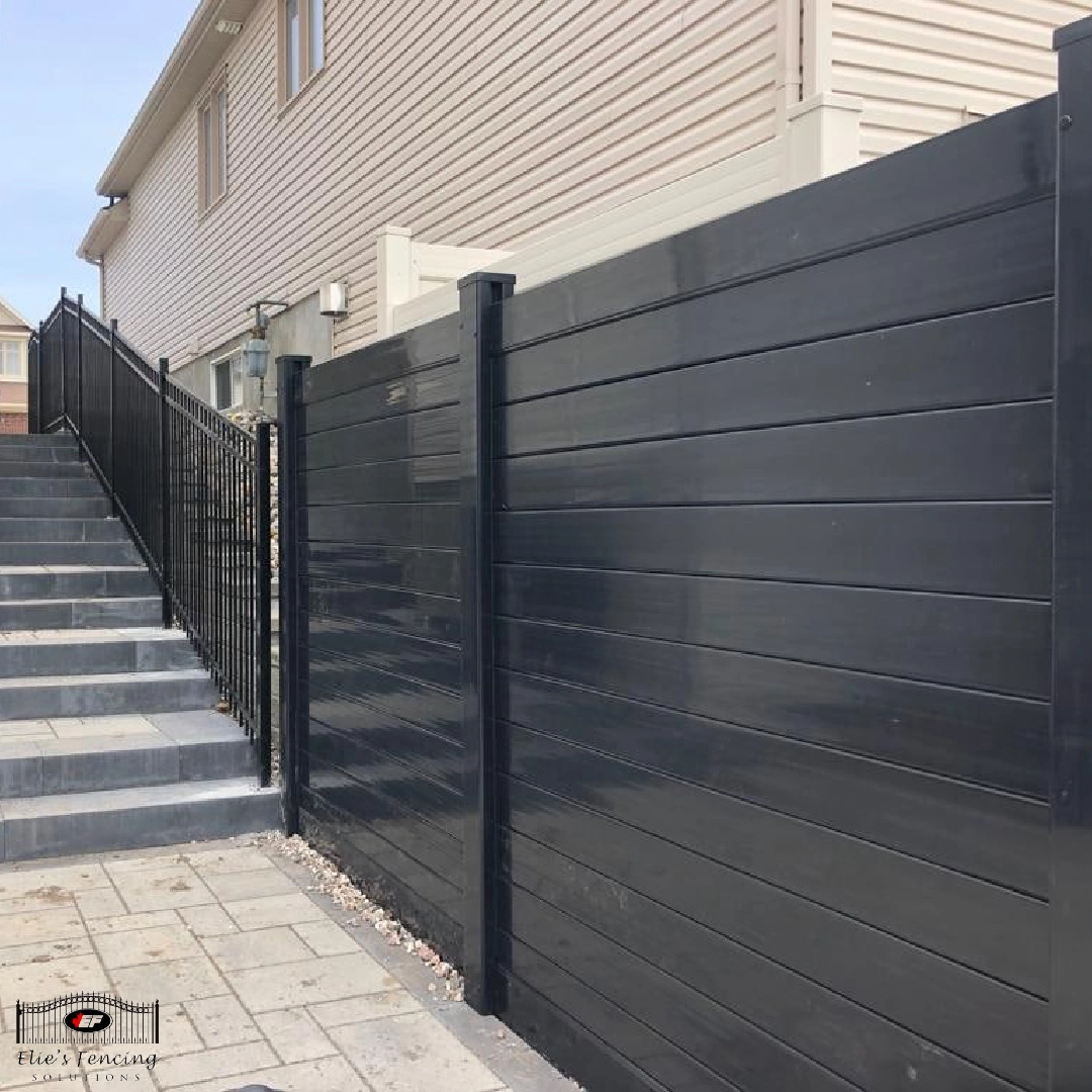 a black fence installed by a skilled fencing contractor borders a path with a stone-tiled surface alongside a beige building with stairs leading up.