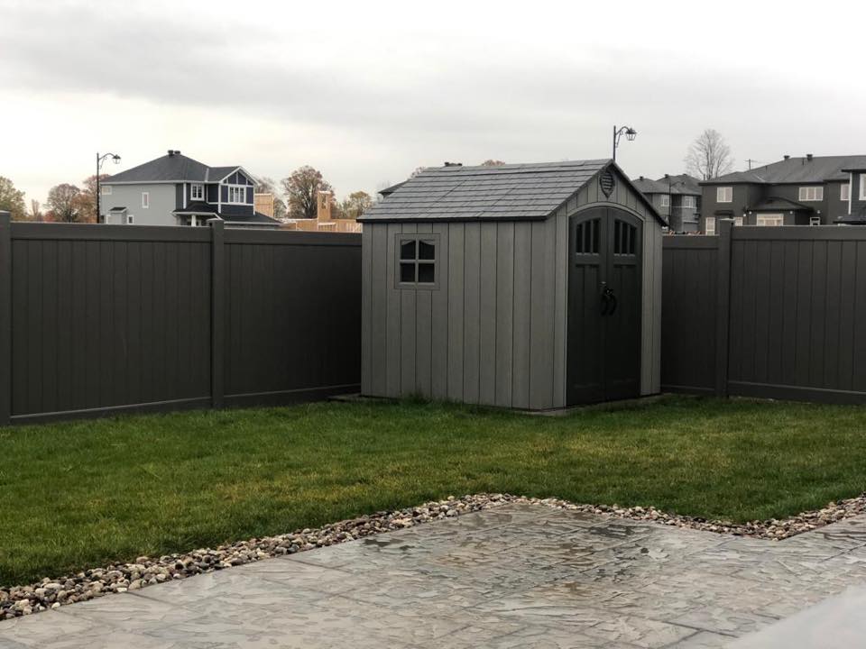 A gray garden shed with a gabled roof stands behind home fences, alongside a green lawn and patterned patio, under an overcast sky.